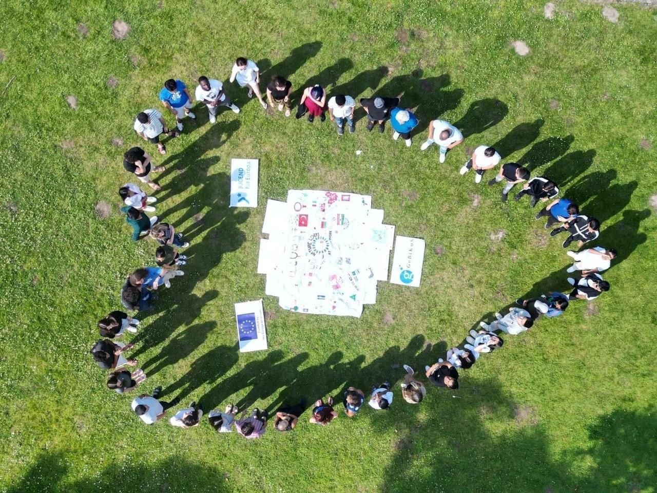 Luftaufnahme einer Gruppe, die auf einer Wiese eine Herzform um Erasmus+- und GeBiKuS-Banner bildet